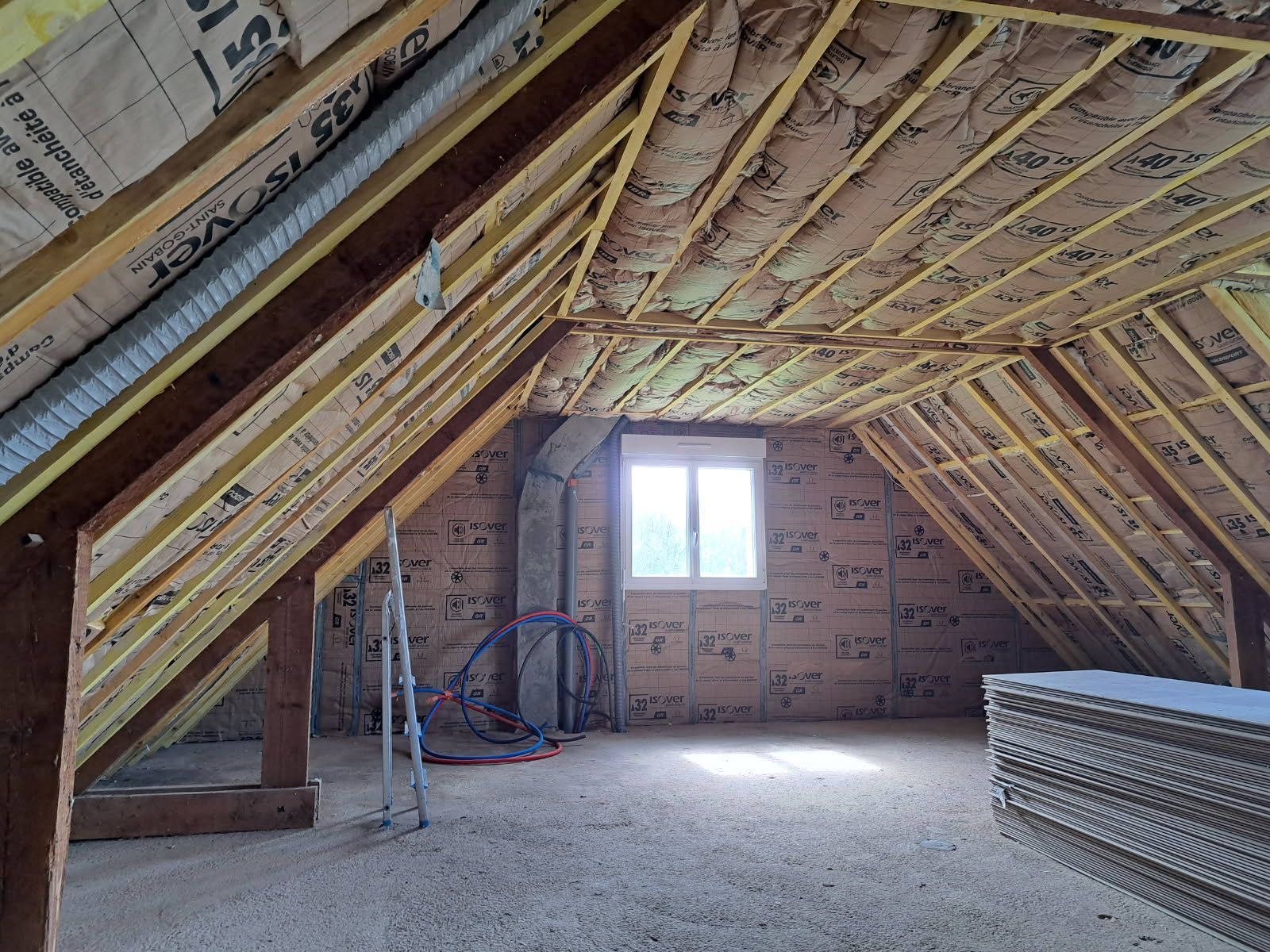 Aménagement intérieur des combles en cours : isolation, poutres apparentes et fenêtre.
