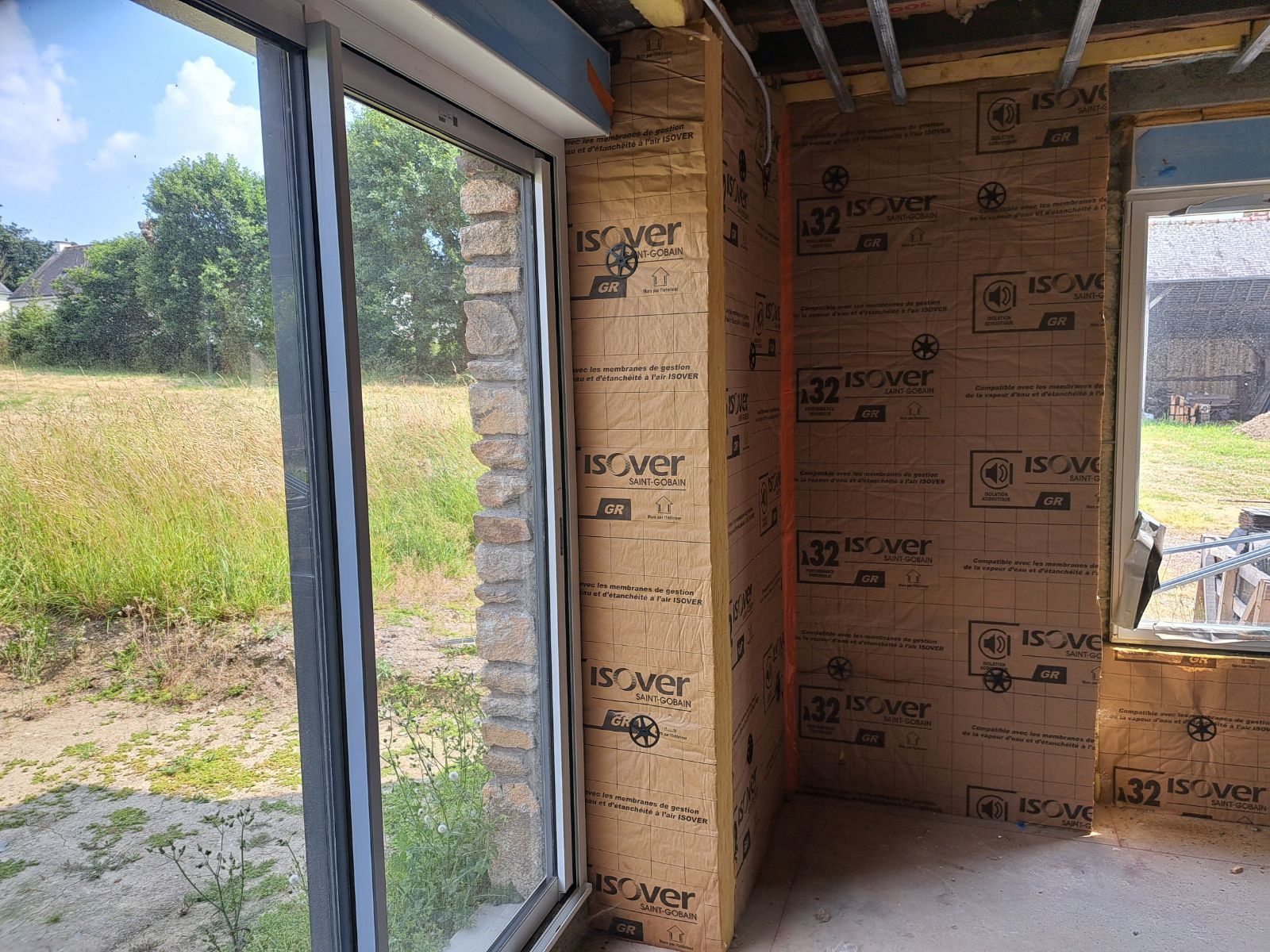 Intérieur d'un bâtiment en construction, montrant des panneaux isolants et une grande fenêtre donnant sur un champ.