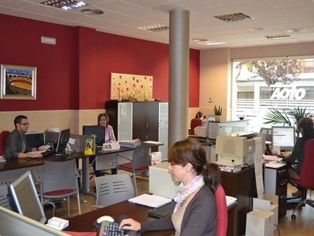 Oficina con varias personas trabajando con computadoras. Pared roja, escritorios y sillas blancos y grises.