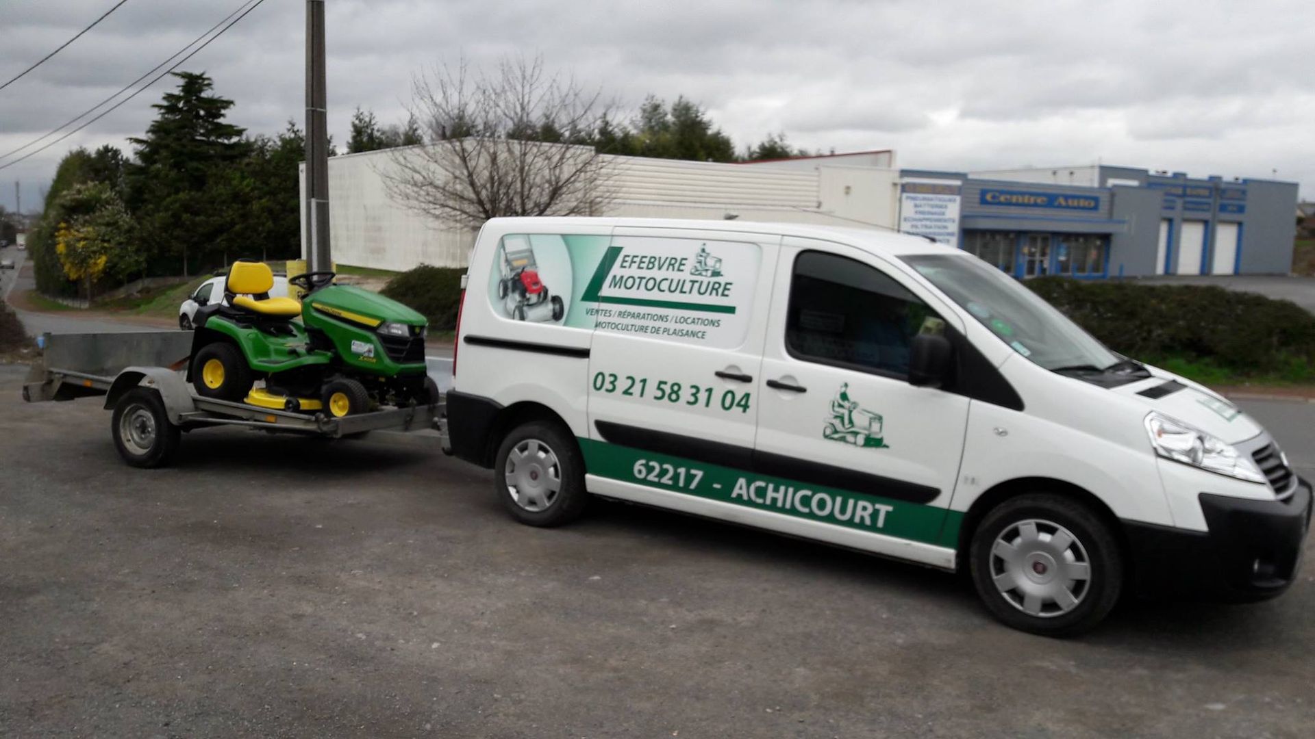 Fourgonnette blanche tractant un tracteur tondeuse sur une remorque ; stationnée à l'extérieur.