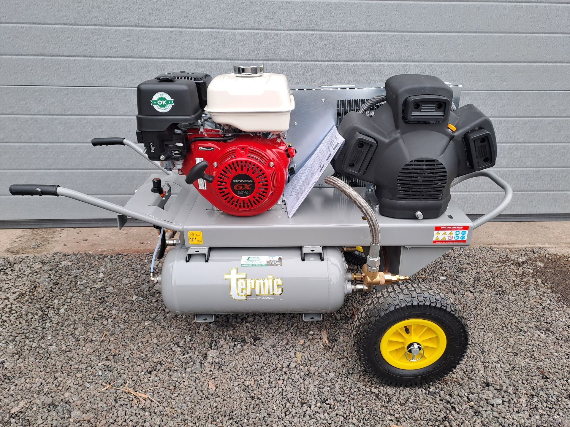 Compresseur d'air gris et rouge avec une petite roue, sur du gravier.