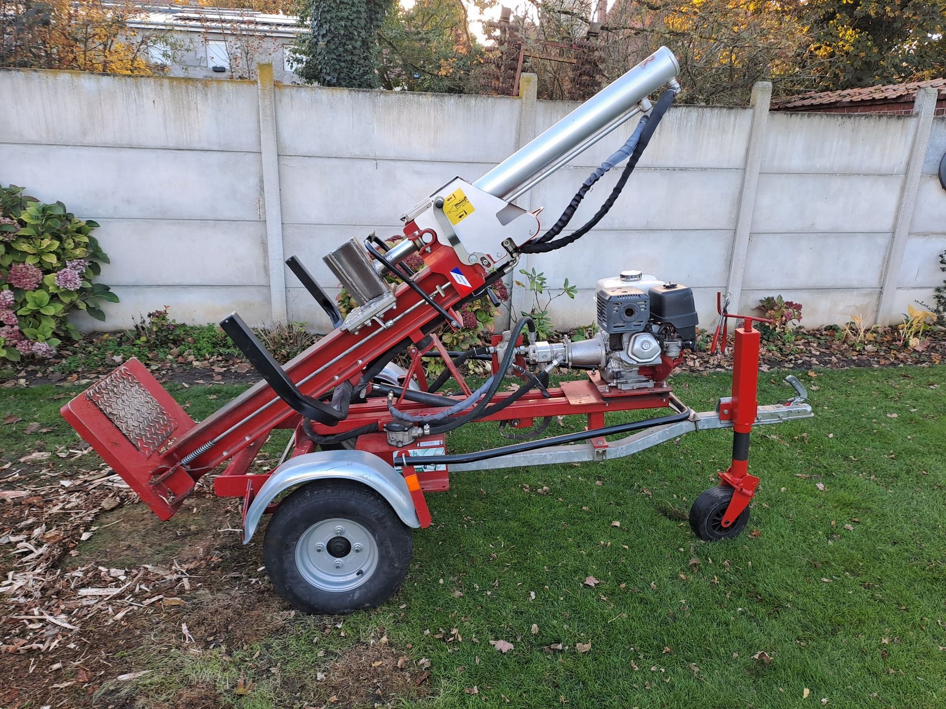 Fendeuse de bûches rouge et argent sur une remorque, dans une cour herbeuse à côté d'un mur en béton.