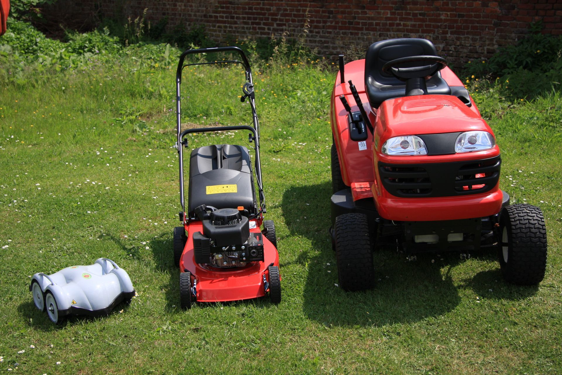 Trois tondeuses à gazon sur une pelouse verte : une tondeuse robotisée, une tondeuse manuelle et une tondeuse autoportée.