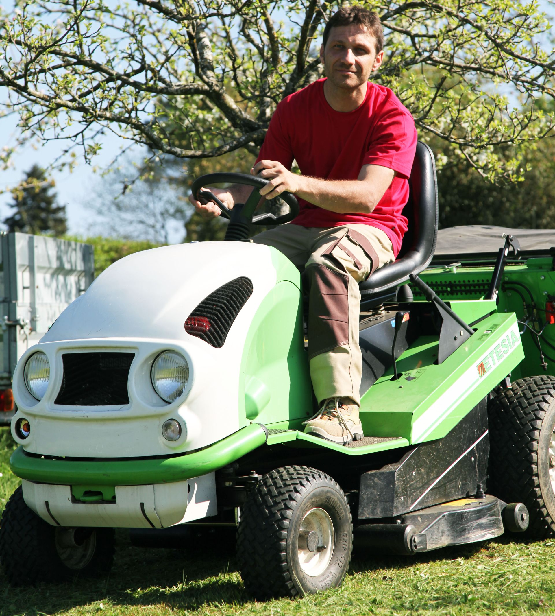 Un homme en chemise rouge sur une tondeuse autoportée blanche et verte dans un jardin.
