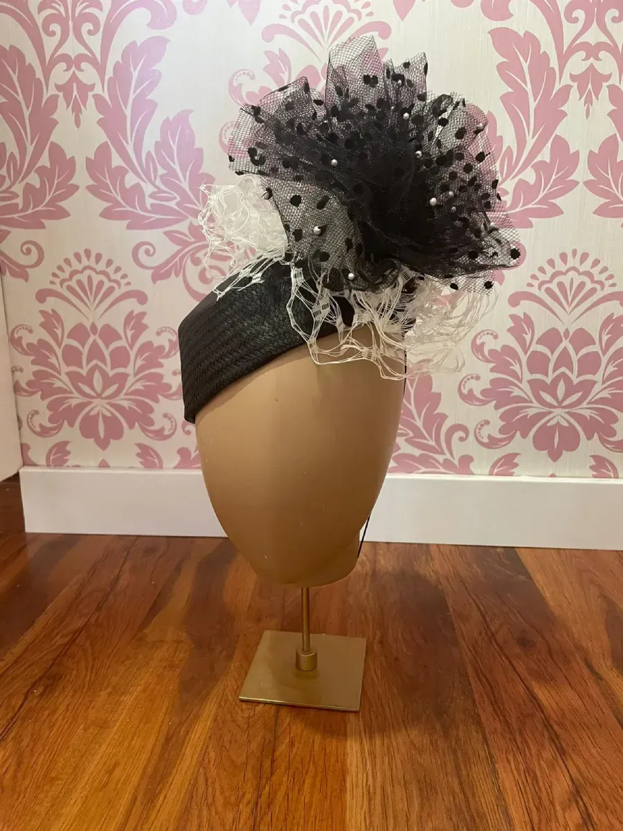 Sombrero formal blanco y negro con red y adornos sobre una cabeza de maniquí, exhibido en un soporte dorado.