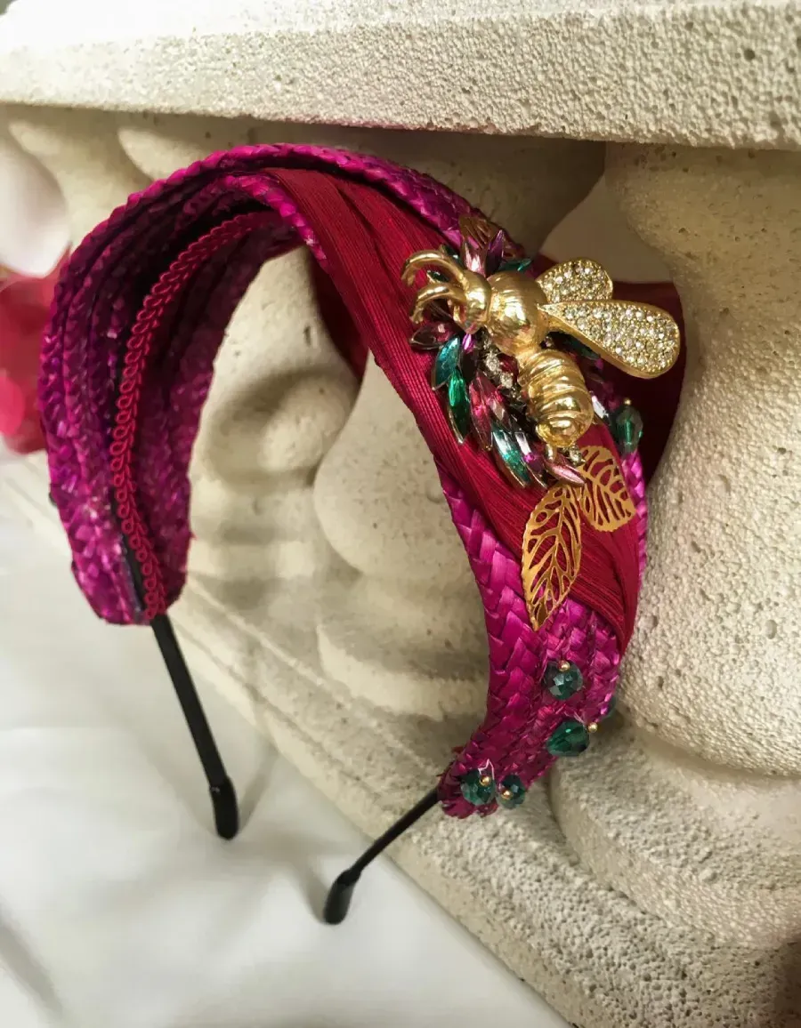 Diadema de cuentas rojas y rosas con una abeja dorada y adornos de joyas, que reposa sobre una columna blanca.