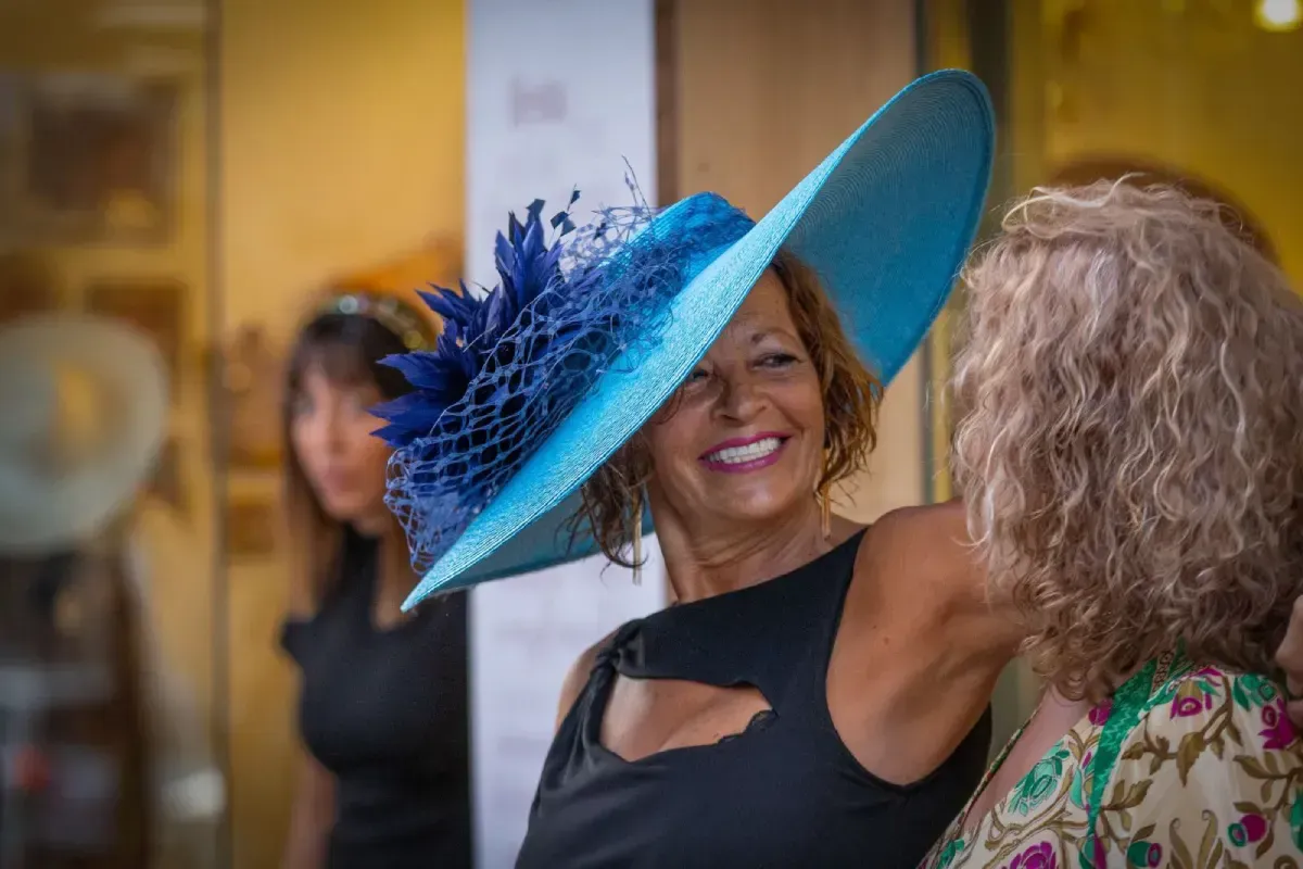 Mujer con sombrero azul con plumas, sonriendo, hablando con otra mujer.