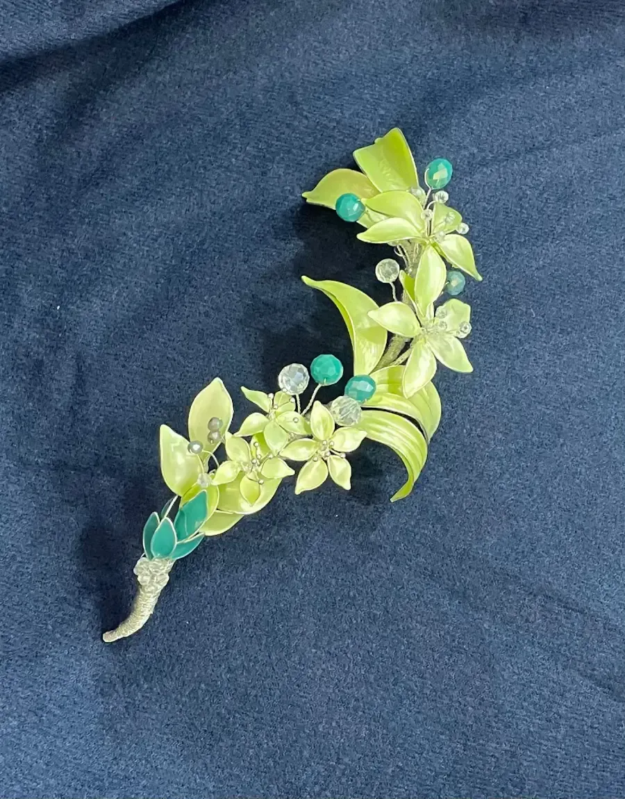 Broche floral verde y plateado con piedras turquesas y transparentes sobre fondo azul marino.