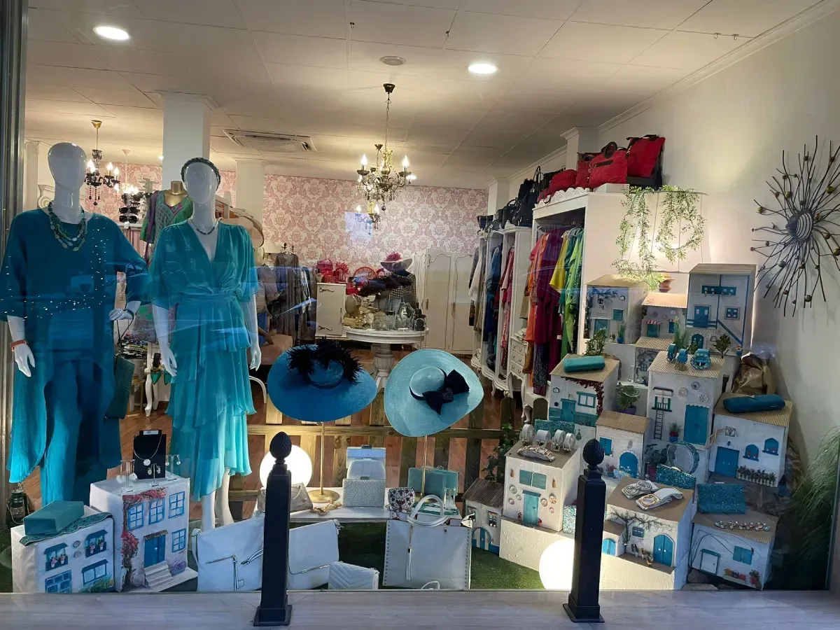 Escaparate de una boutique con vestidos turquesas sobre maniquíes y casas decorativas azules y blancas.