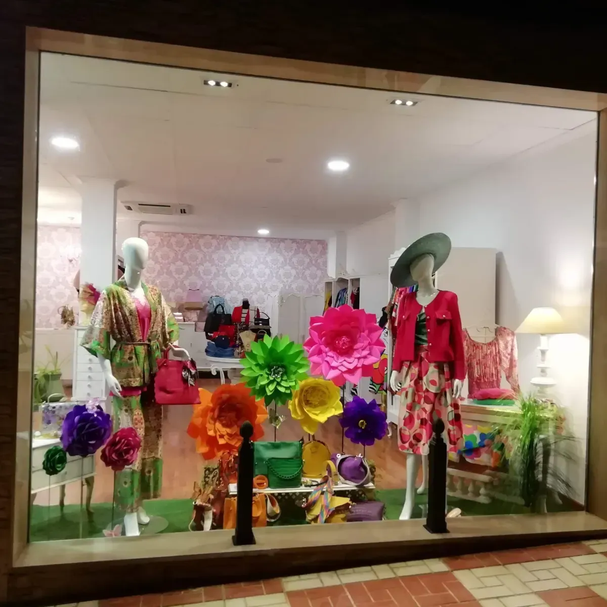 Exhibición en escaparate con maniquíes luciendo trajes coloridos, rodeados de grandes flores de papel y bolsos.