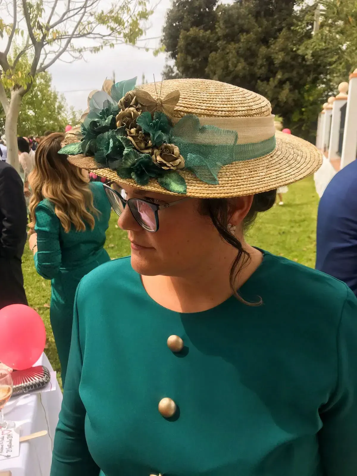 Mujer con vestido verde y sombrero de paja con decoraciones florales al aire libre, cerca de una fiesta.