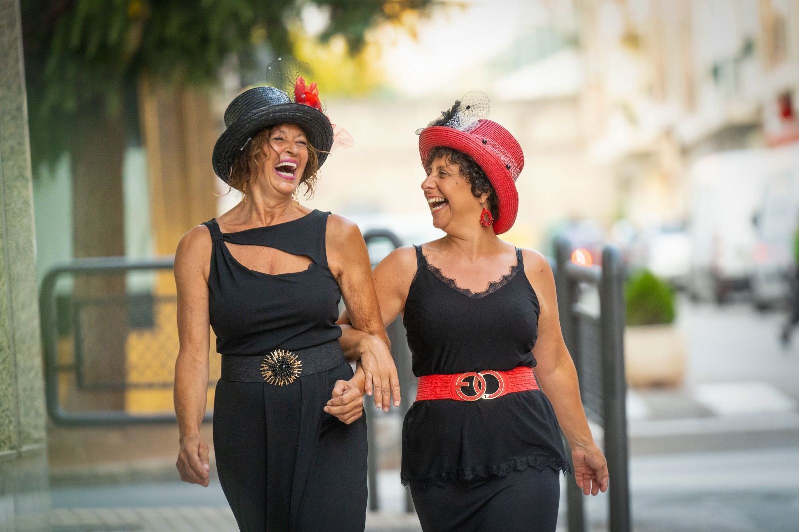 Dos mujeres con sombreros, riendo y caminando del brazo por una acera.