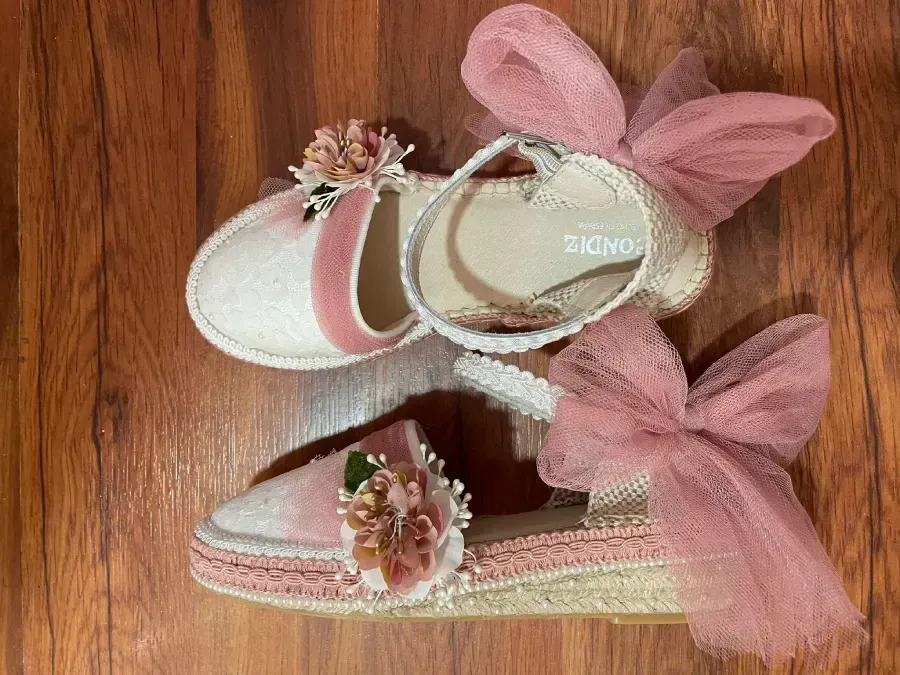 Dos pares de zapatos de bebé de color rosa y blanco con detalles florales y lazos, sobre superficie de madera.
