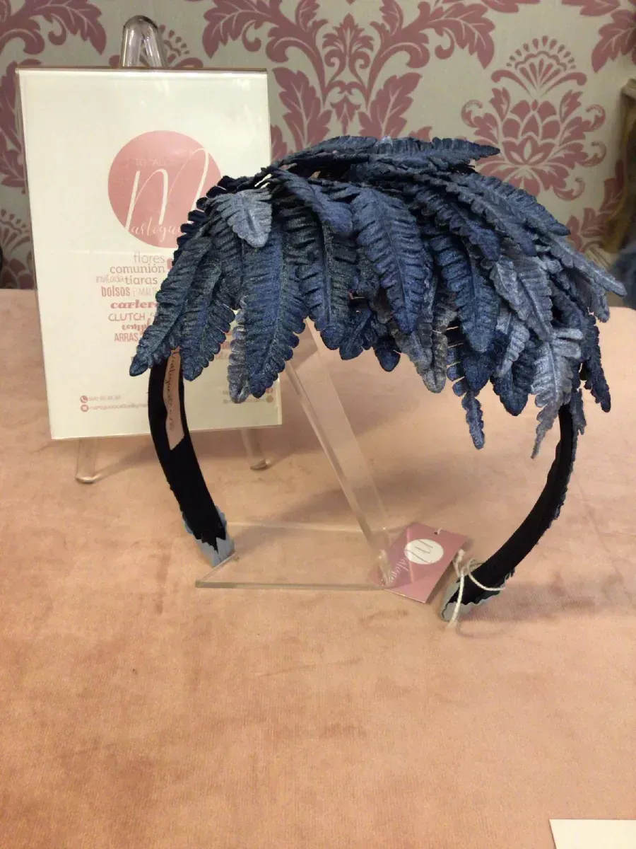 Diadema de plumas azules en exhibición, sobre una superficie rosa y con un cartel en el fondo.