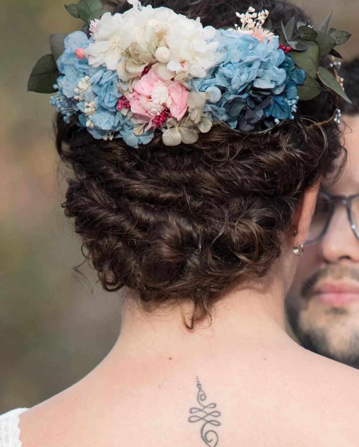 Persona con corona de flores y peinado recogido, de espaldas, con un pequeño tatuaje en el cuello.