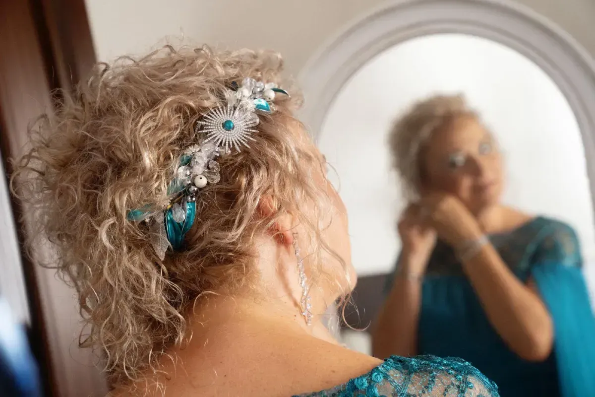 Mujer con cabello rubio rizado que lleva un peluquín verde azulado, mirándose en un espejo y ajustándose aretes.