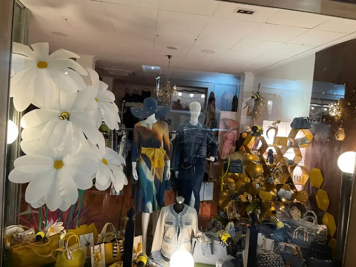 Escaparate de tienda con maniquíes con ropa colorida, grandes decoraciones florales blancas y acentos amarillos.