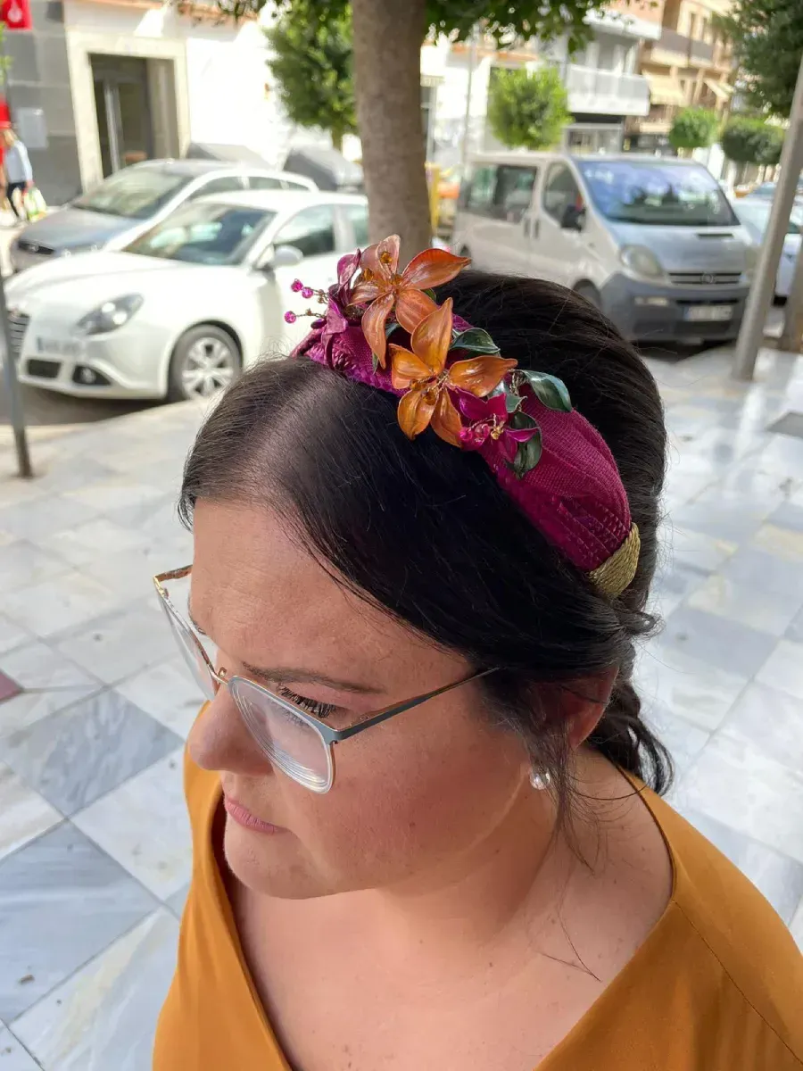 Mujer con gafas y una diadema color burdeos con flores naranjas, de pie afuera.