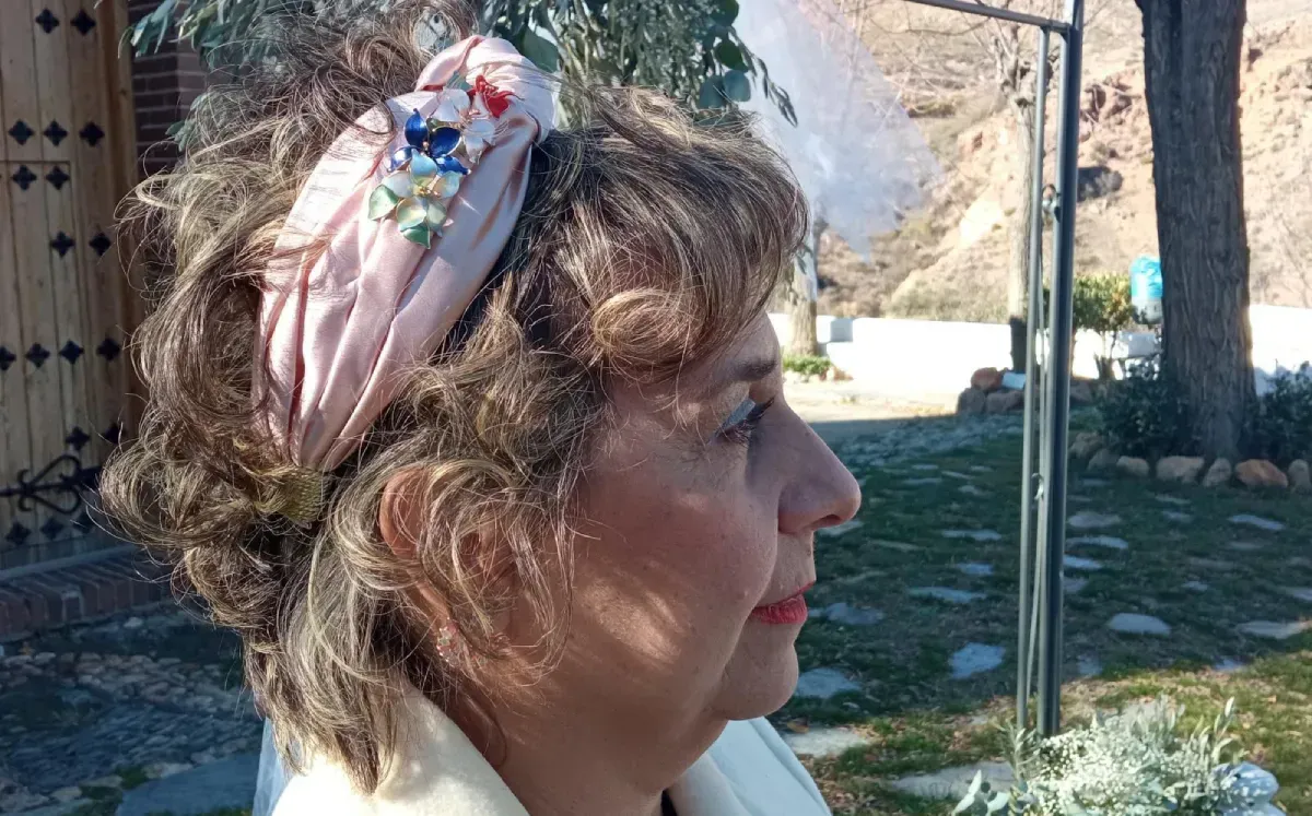 Mujer que lleva un pañuelo floral rosa al aire libre, mirando hacia la derecha, con árboles y una montaña de fondo.