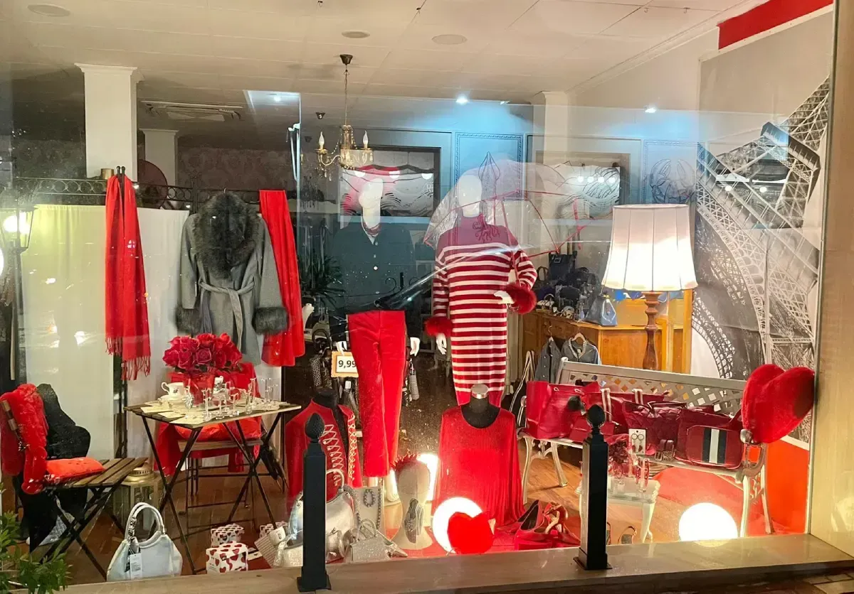 Escaparate con ropa y adornos rojos, entre maniquíes, bufandas y mesa.