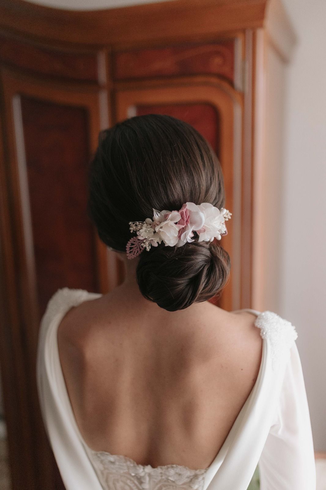 Mujer con un vestido de novia blanco, cabello recogido en un moño con un tocado floral, de pie cerca de un armario de madera.