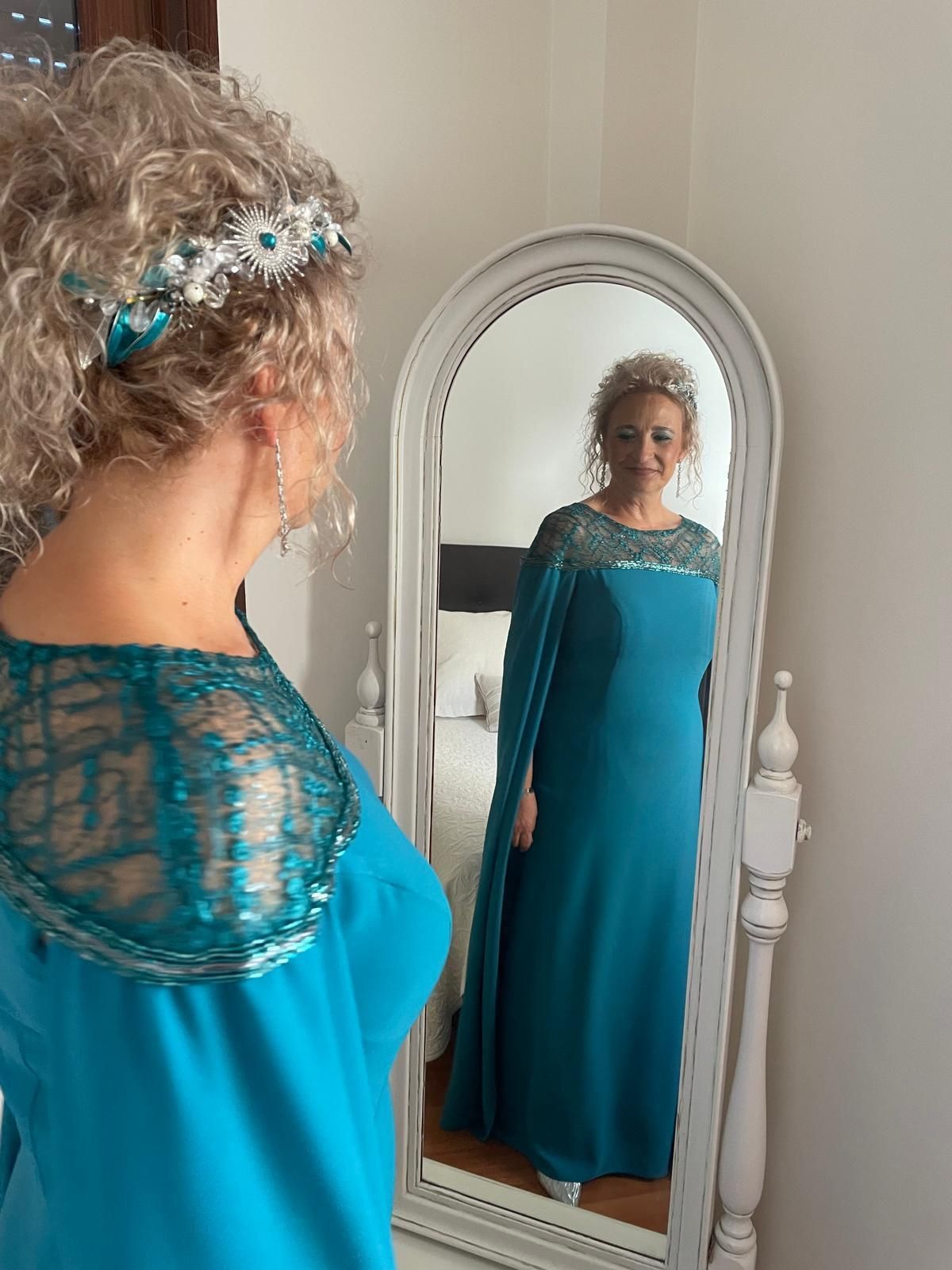 Mujer con vestido verde azulado y capa mirándose en un espejo. Tiene el pelo rizado y una diadema con joyas.