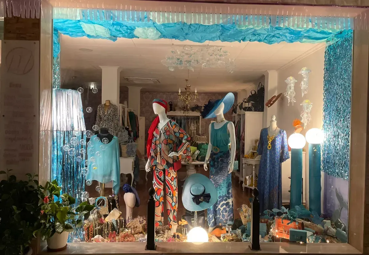 Escaparate: maniquíes con ropa de colores, decoraciones azules y turquesas, luces brillantes.