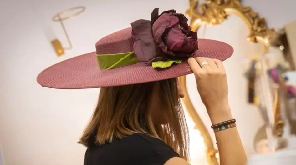 Mujer con un sombrero rosa y una flor grande. Está en una habitación con un espejo dorado.