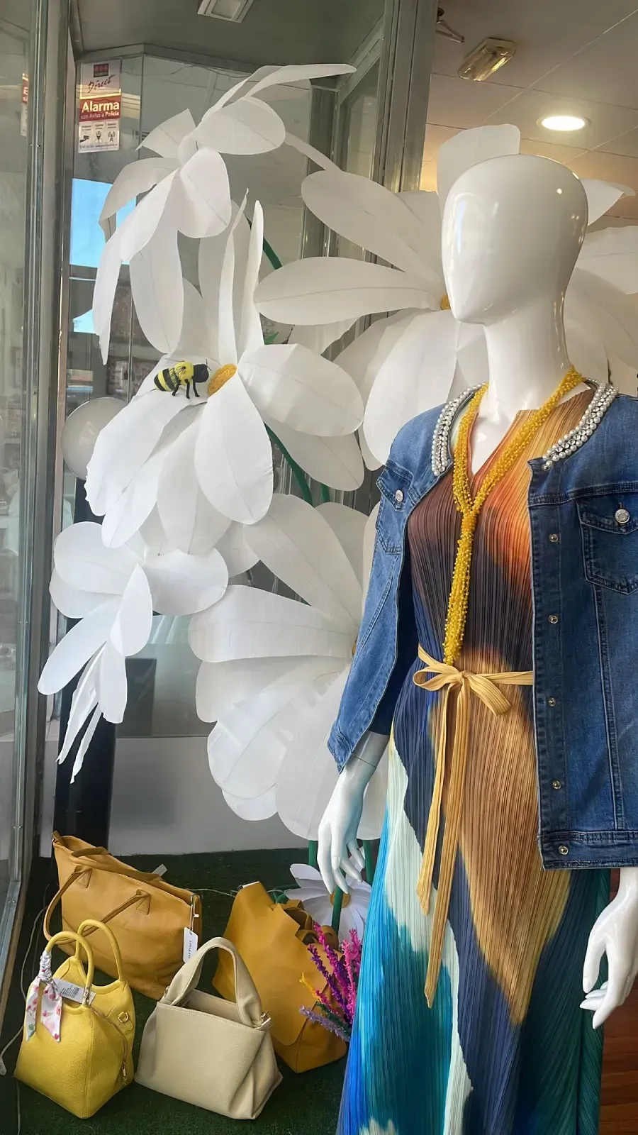 Maniquí en el escaparate de una tienda con un vestido colorido y una chaqueta vaquera, rodeado de flores gigantes.