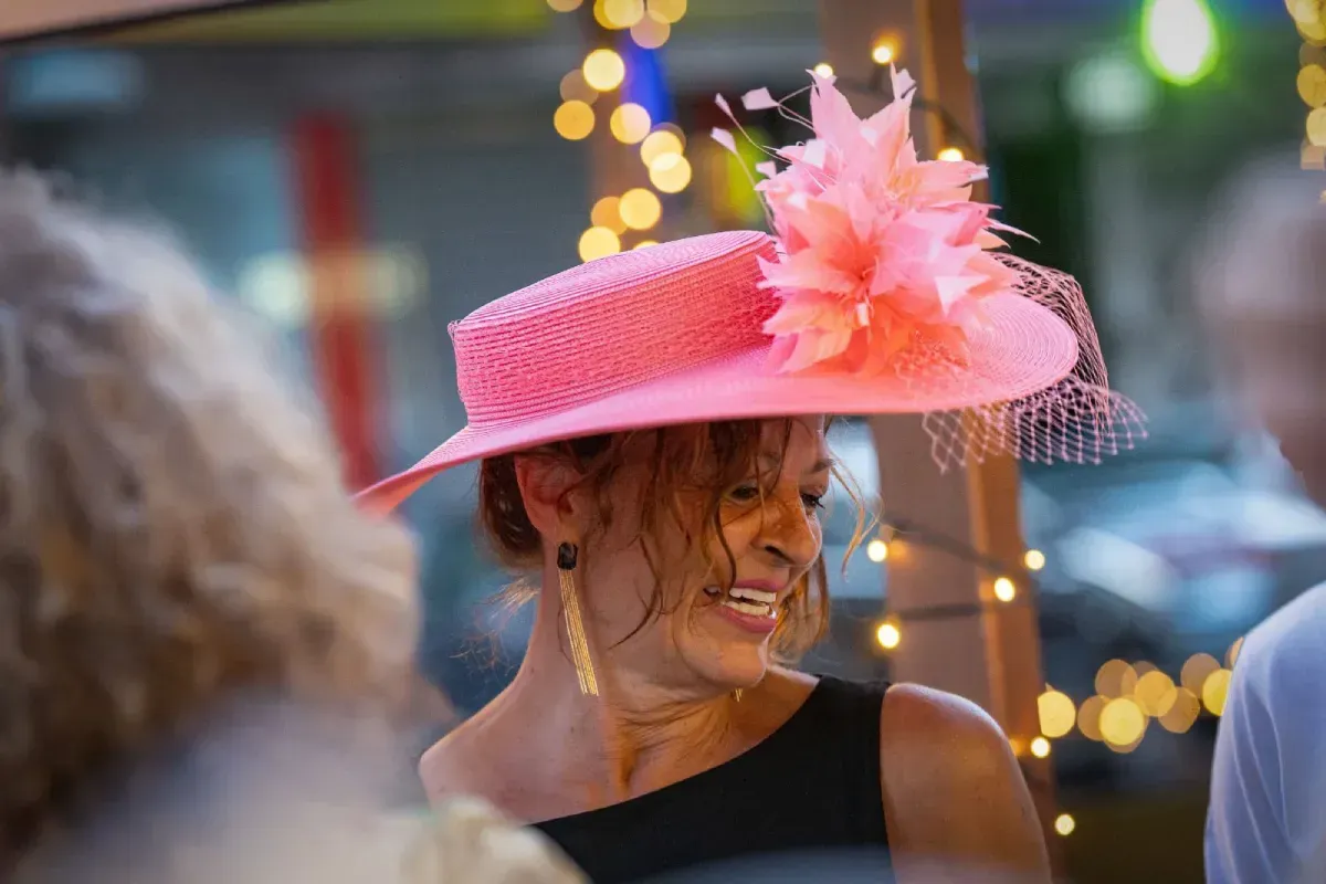 Mujer sonriendo, con un sombrero rosa con plumas y red, en un evento al aire libre con luces de cadena.
