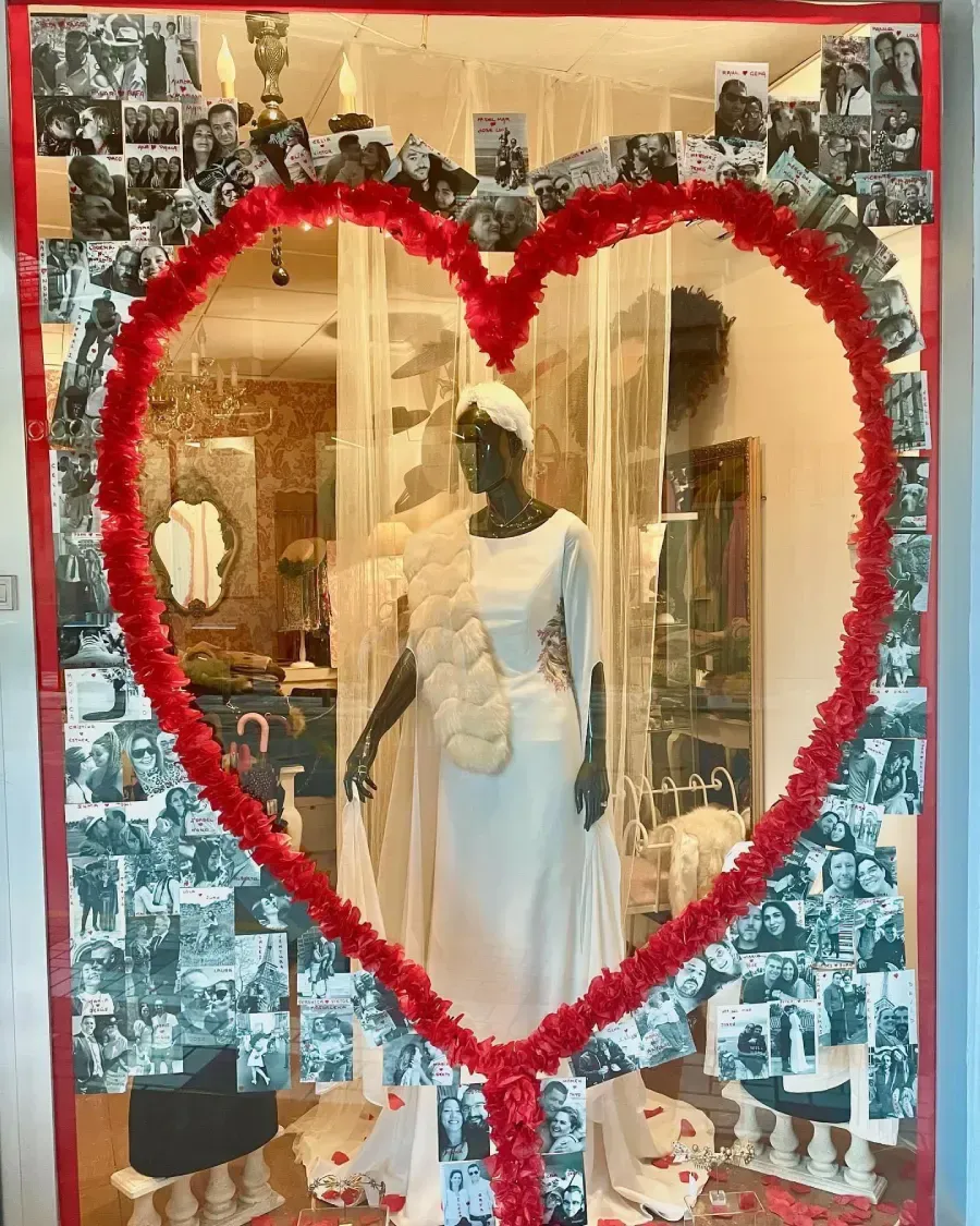 Maniquí con vestido de novia blanco en un escaparate, enmarcado por un corazón rojo y fotografías vintage.