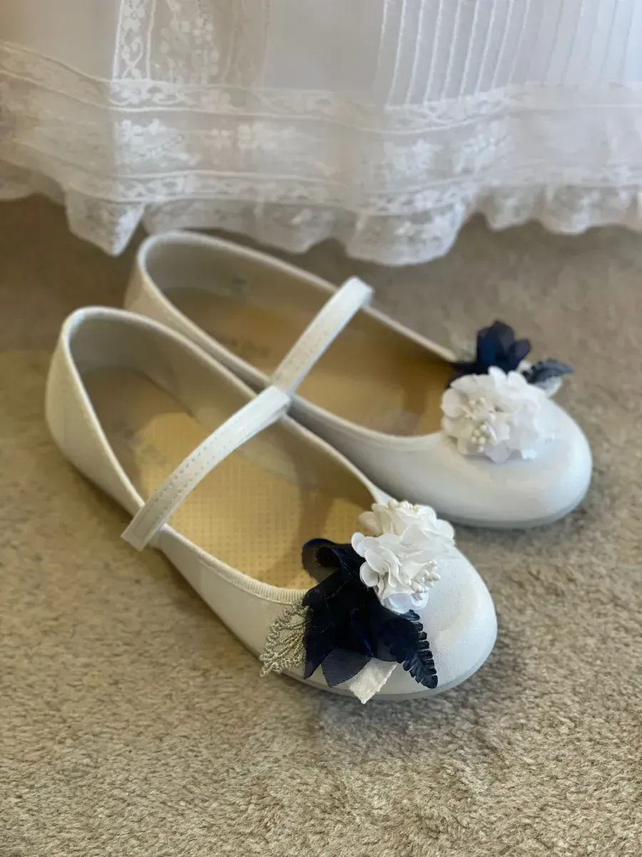 Zapatos de vestir blancos con detalles florales, sobre una superficie beige cerca de una tela blanca.