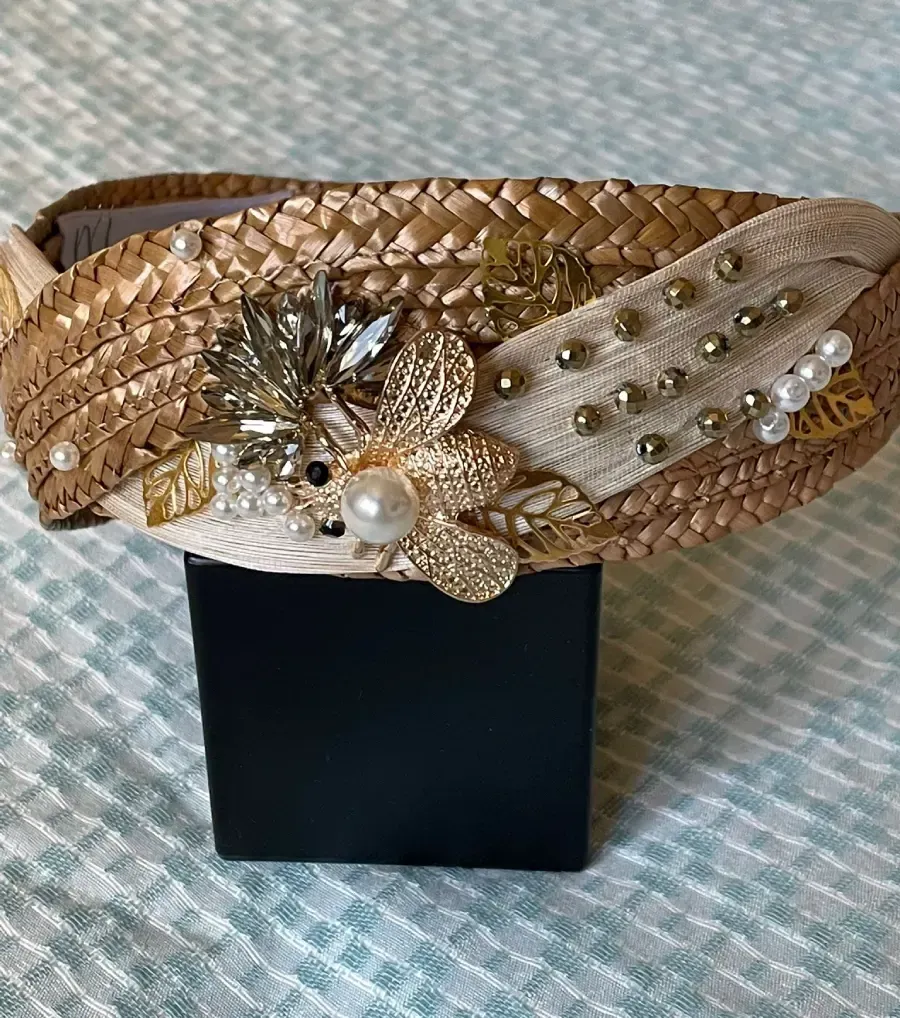 Diadema tejida en dorado y beige con adornos florales y de cuentas, exhibida sobre un soporte negro.