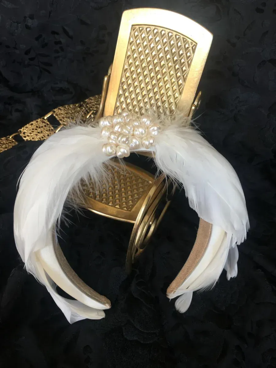 Diadema estilo Art Decó en tono dorado con plumas blancas y adorno de perlas, sobre fondo negro.