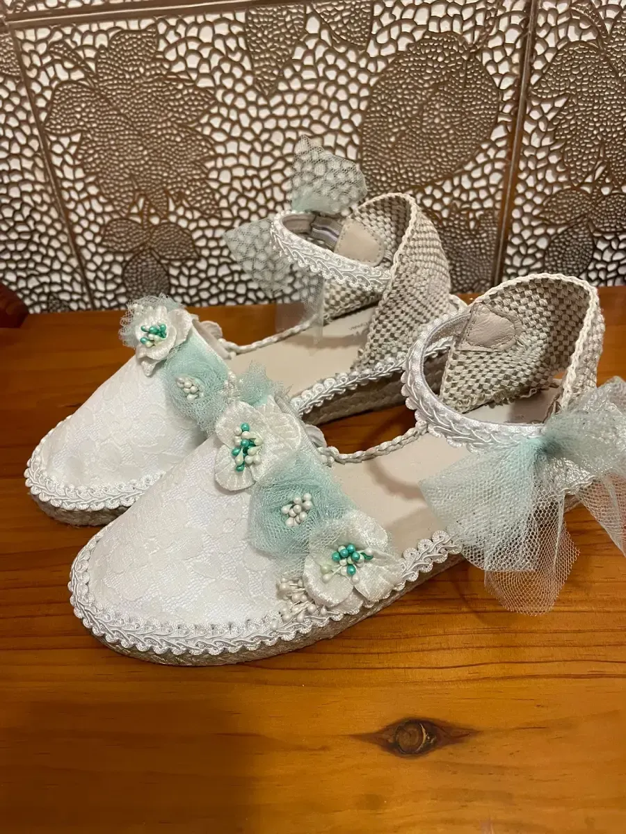 Zapatos de vestir de color blanco y plata con detalles florales y de encaje sobre superficie de madera.