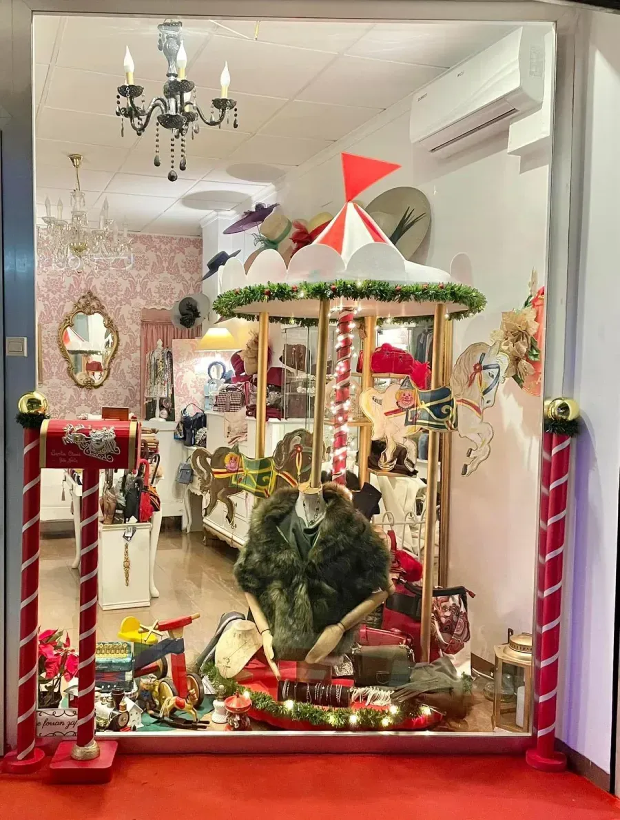 Escaparate con carrusel, sombreros y adornos navideños. El rojo, el dorado y el verde dominan la escena.