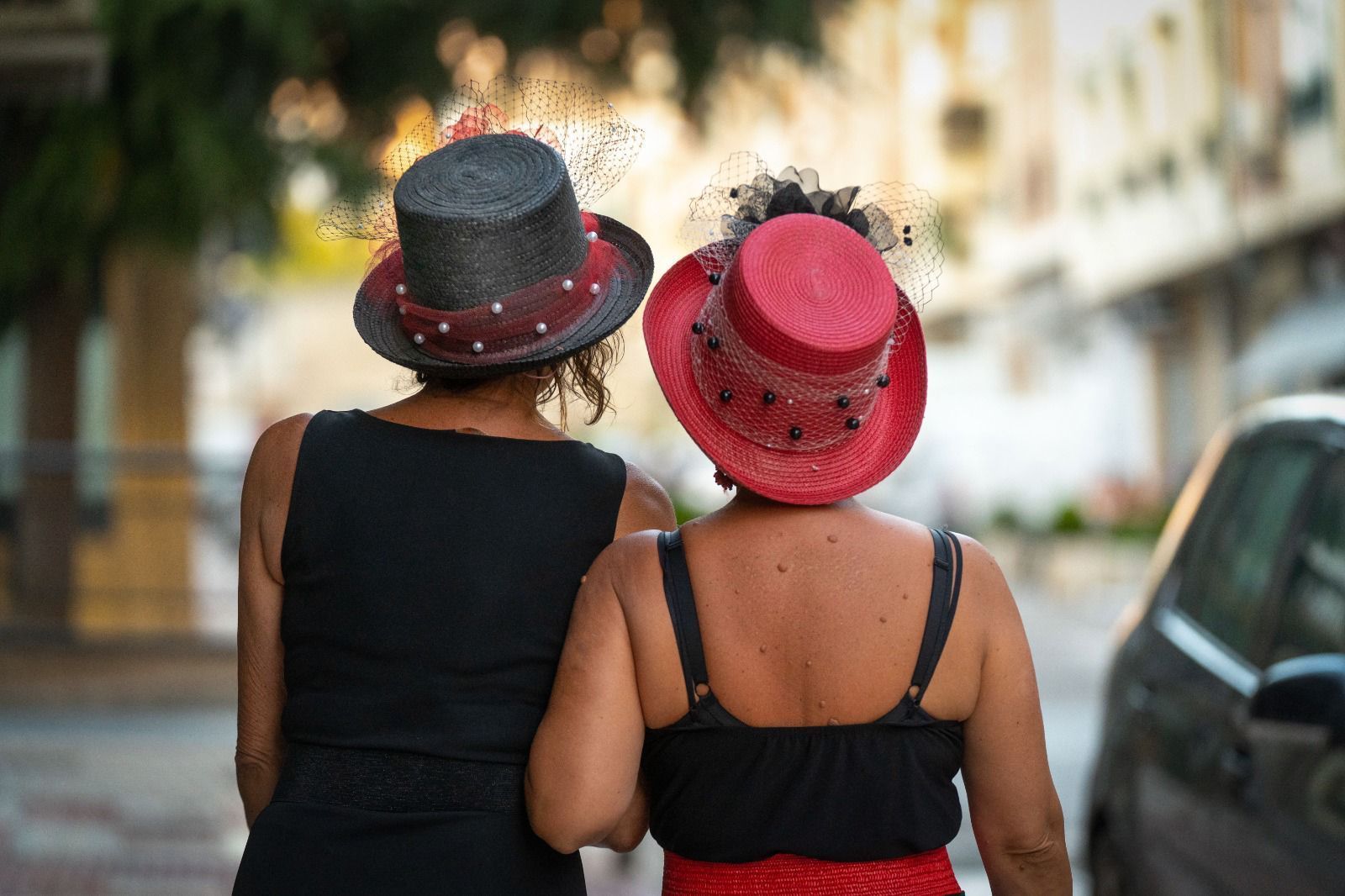 Dos mujeres caminando con sombreros decorativos negros y rojos. Fondo: calle de la ciudad.