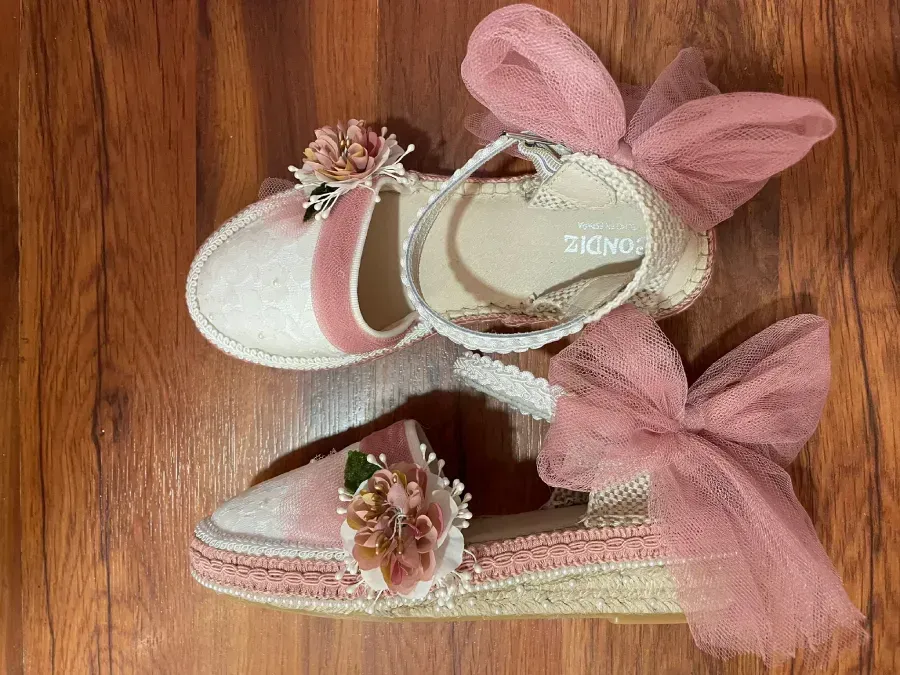 Zapatos de bebé rosas y blancos con detalles florales y grandes lazos de tul, sobre un piso de madera.