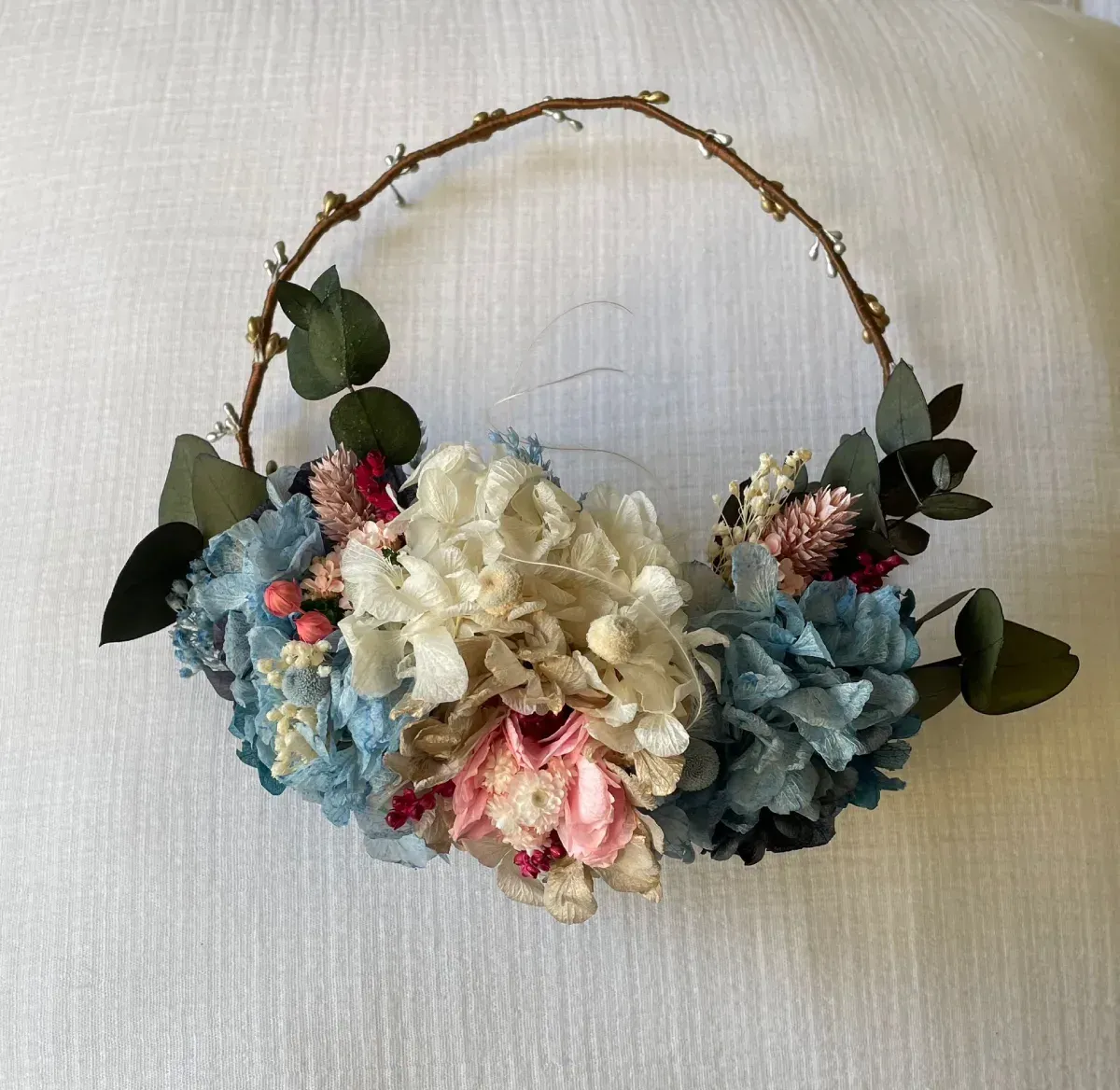 Corona floral con flores azules, rosas y crema, hojas verdes y un fino anillo de enredadera marrón.