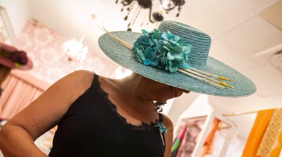Mujer con un sombrero azul de ala ancha con detalles florales verde azulado, mirando hacia abajo en una tienda.