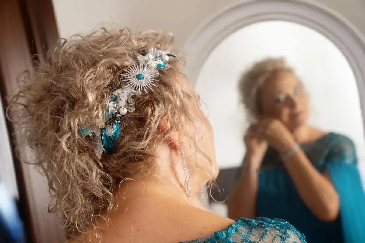 Mujer con cabello rizado y tocado decorativo, mirándose al espejo y tocándose el pendiente. Top turquesa.