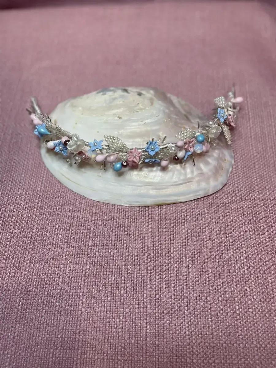 Delicada diadema floral con cuentas rosas, azules y blancas sobre una concha, sobre un fondo texturizado rosa.