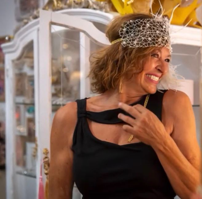 Mujer sonriente, vistiendo un top negro, tocado con plumas, delante de un mueble blanco.