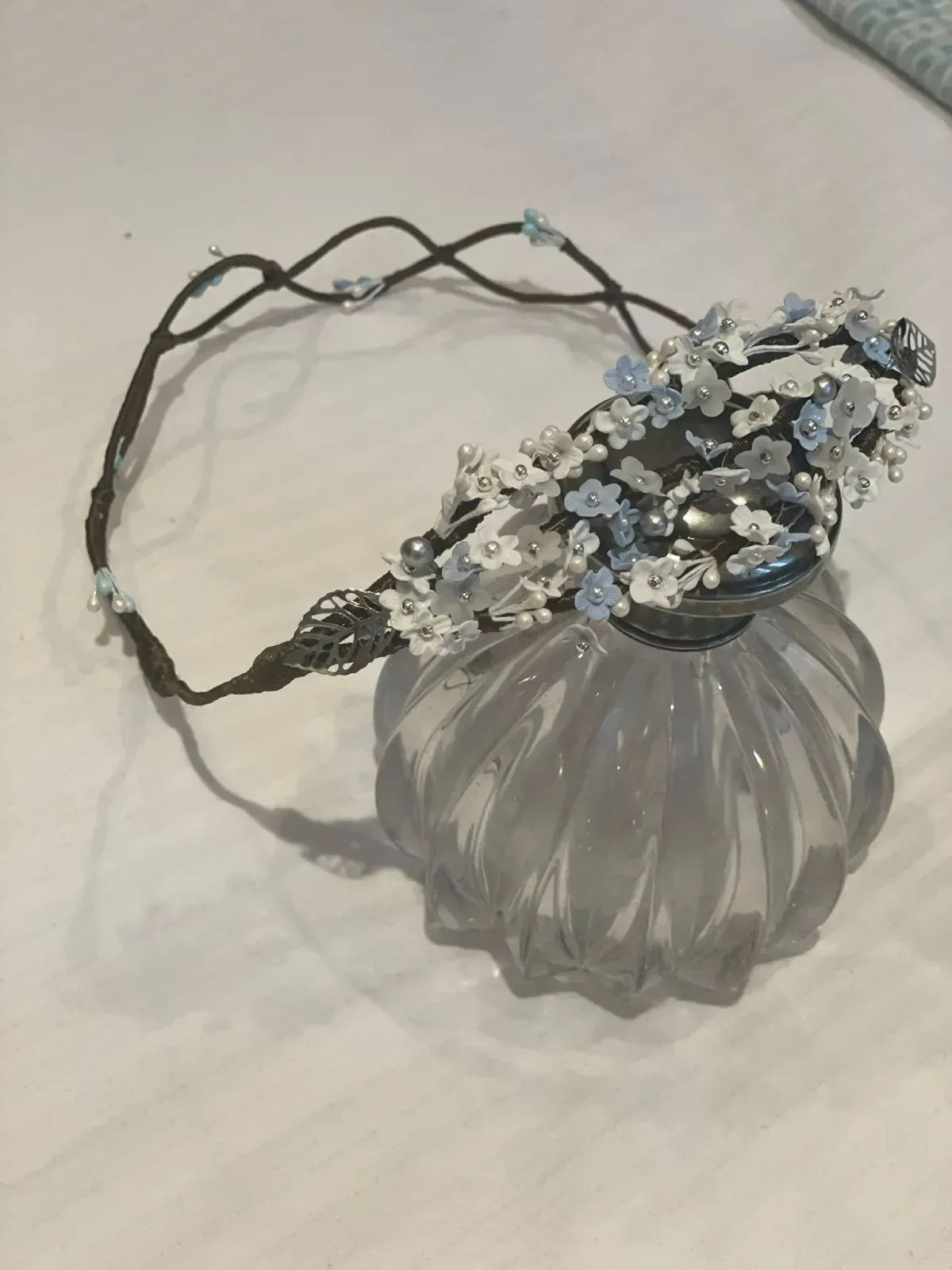 Una corona floral con flores blancas y azules reposa sobre un frasco de perfume de vidrio decorativo.