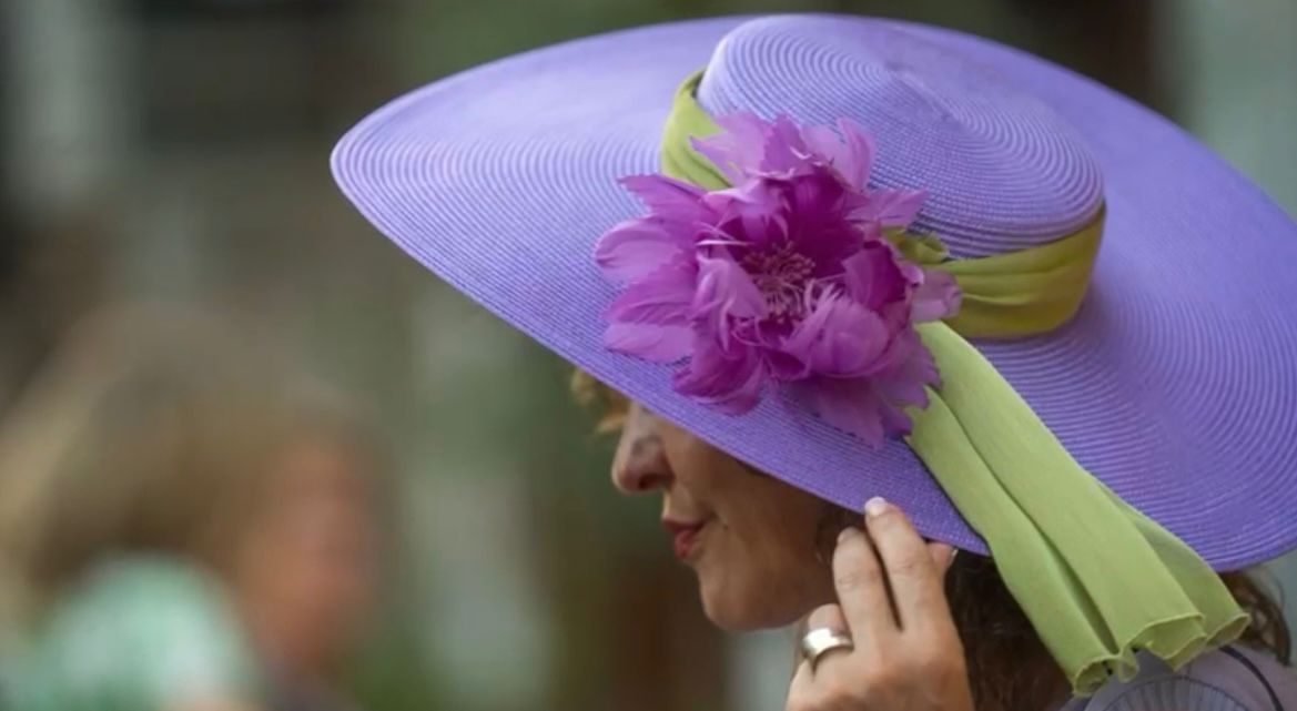 Mujer de perfil con un gran sombrero lavanda con una flor violeta y una cinta verde.
