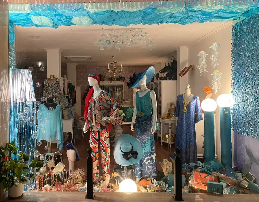 Escaparate con maniquíes vestidos con ropa colorida, adornados con adornos azules y blancos y luces brillantes.