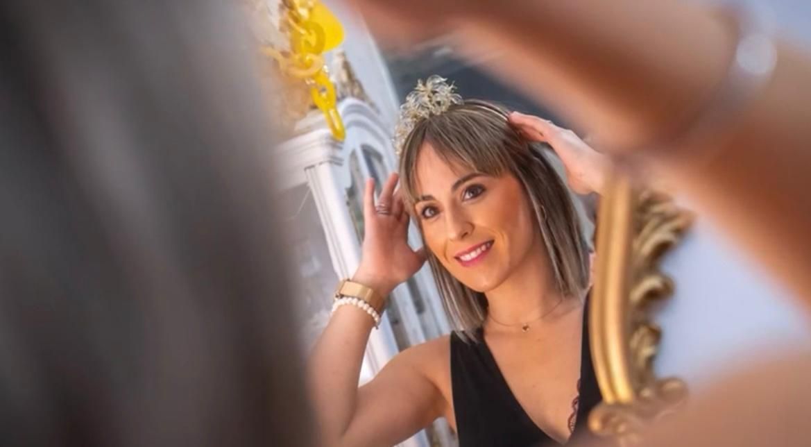 Una mujer se mira al espejo ajustándose una tiara. Sonríe, con un vestido negro.