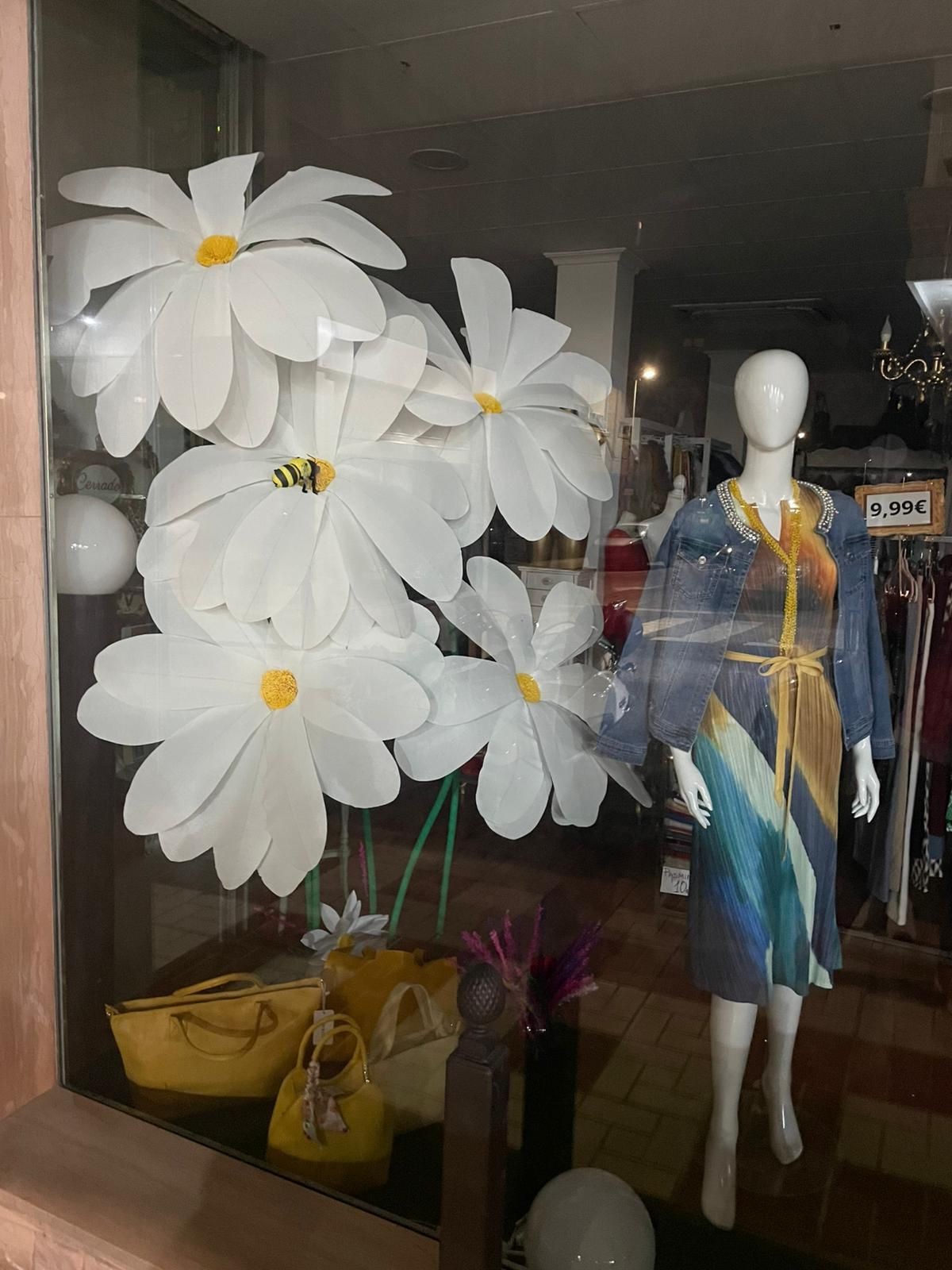 Escaparate: margaritas blancas, maniquí con vestido azul, bolsos amarillos.