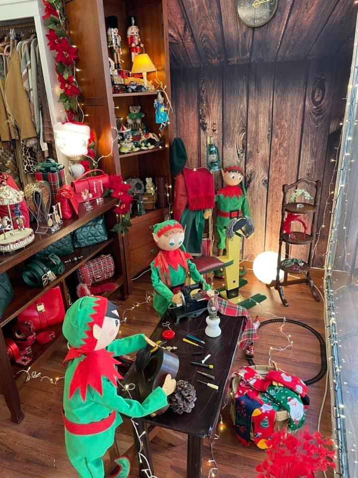 Exhibición navideña con figuras de elfos trabajando en un taller, fondo de madera, estantes con decoración.