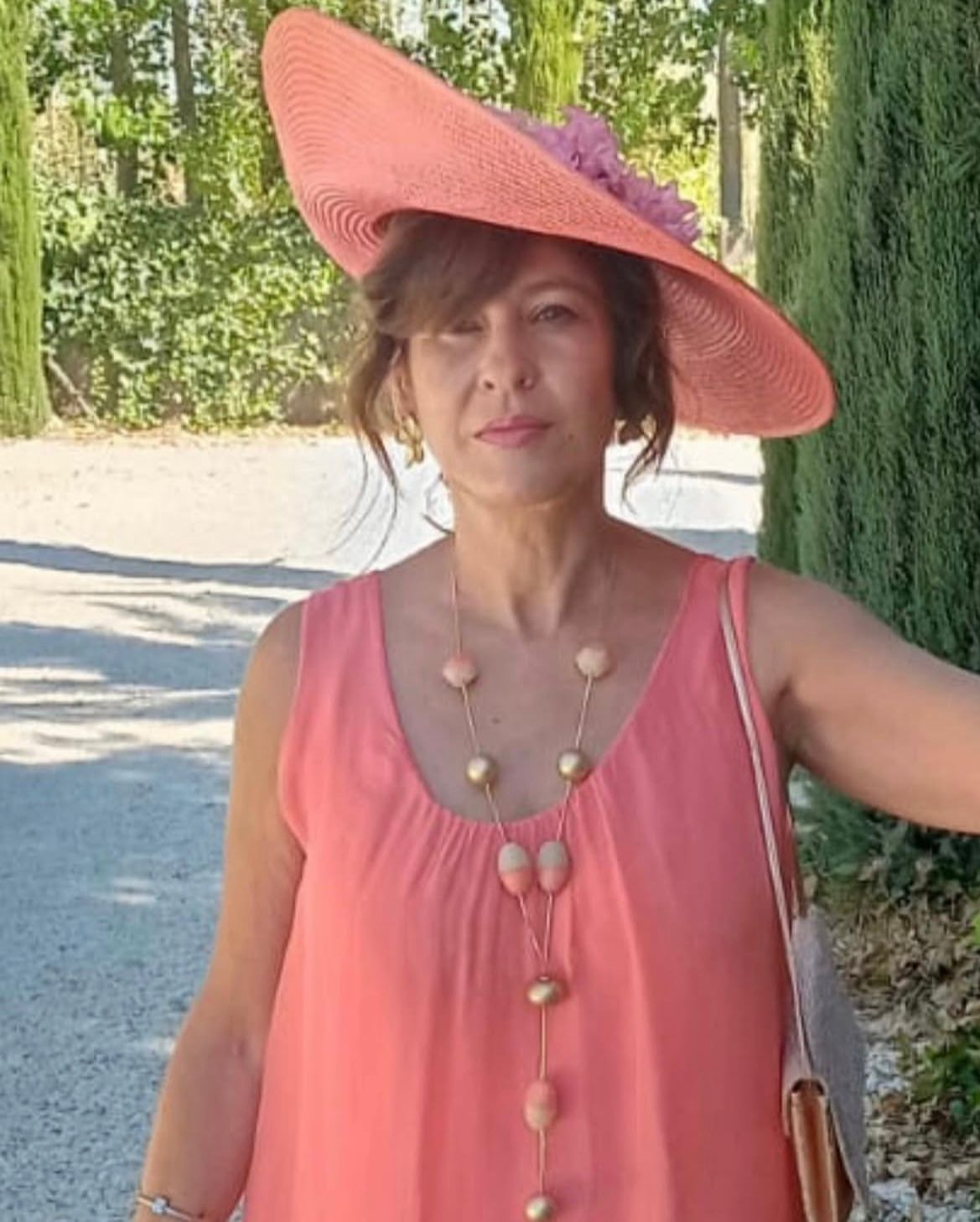 Mujer con vestido y sombrero color melocotón al aire libre, con un collar largo; árboles en el fondo.